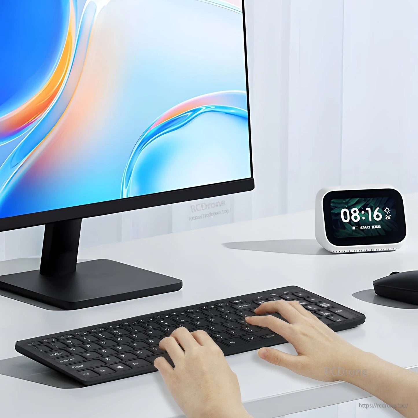 Wireless keyboard in use on desk with monitor and smart clock, hands typing with mouse nearby