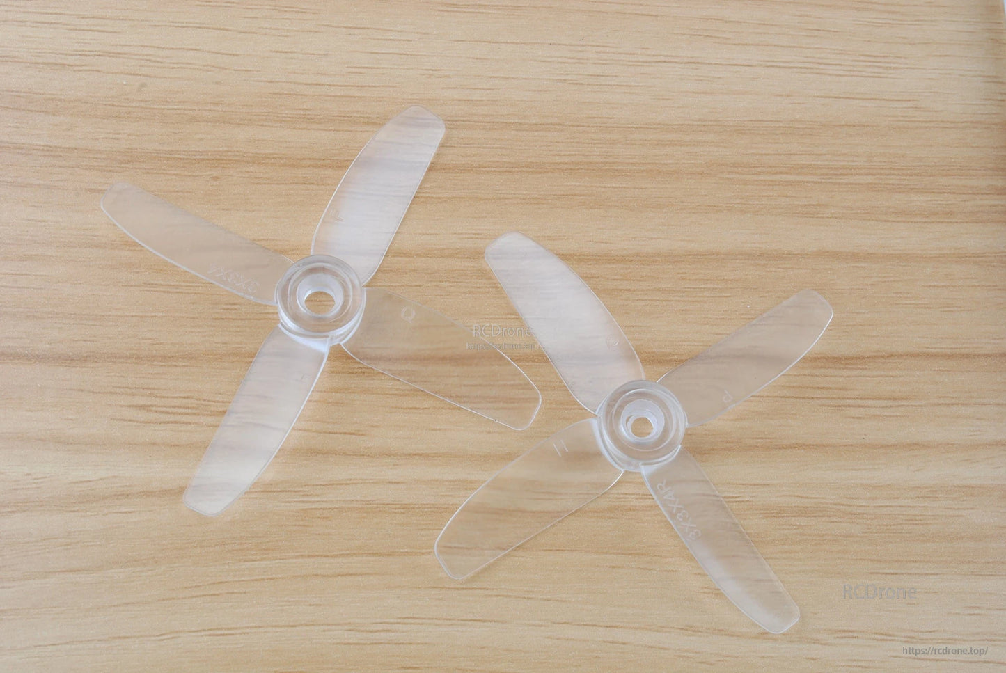 Clear translucent 4-blade propellers pair on wooden surface, top-down view showing hubs and blade shape