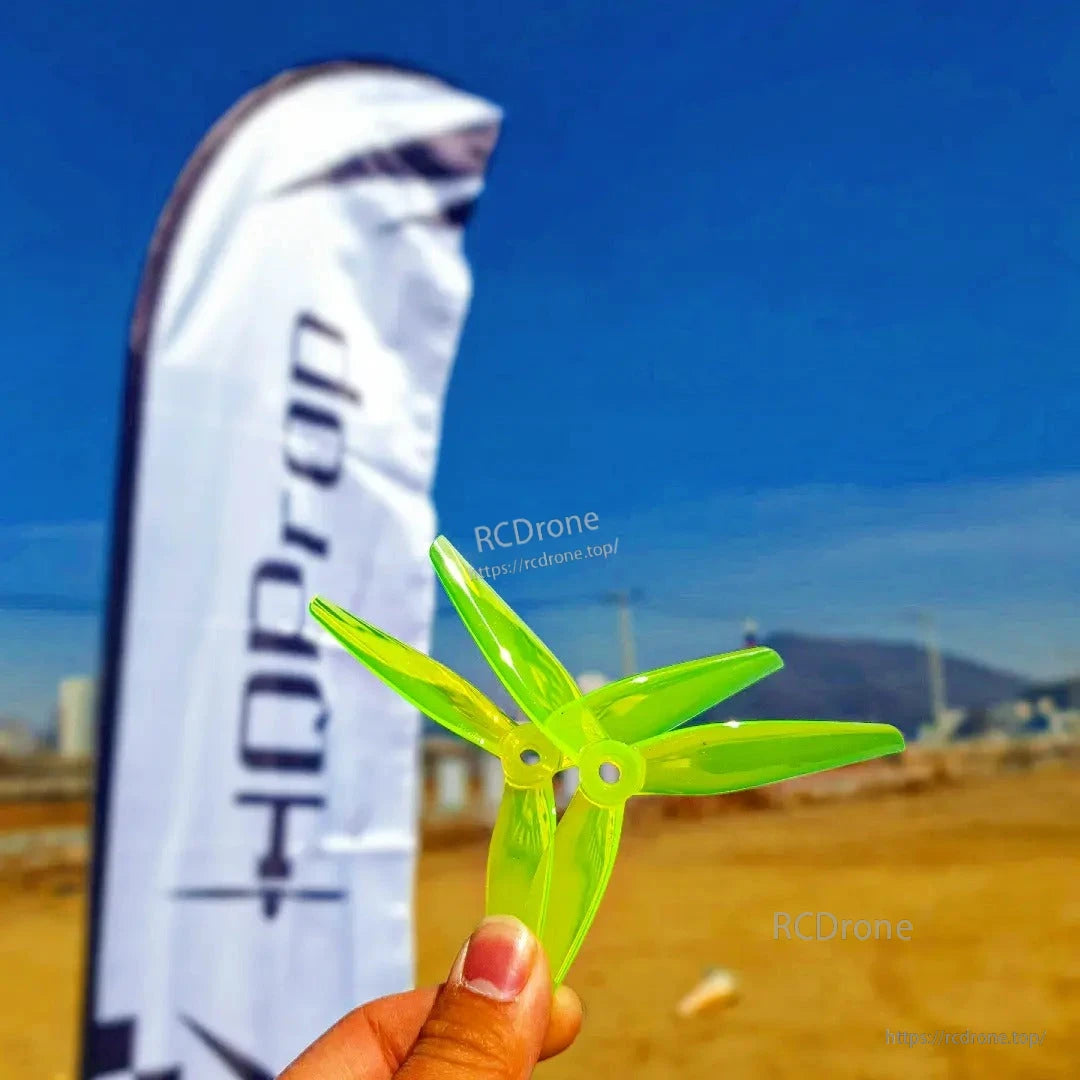 Hand holding a stack of neon green tri-blade propellers outdoors with HQProp flag in background