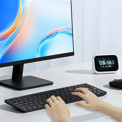 Wireless keyboard in use on desk with monitor and smart clock, hands typing with mouse nearby