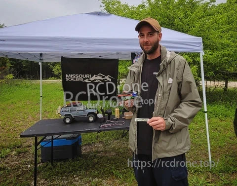 Outdoor event setup under a canopy with an RC vehicle on a table and a person holding a small product box