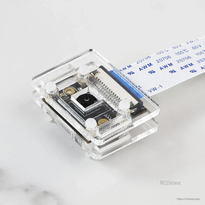 Top view of camera module in clear acrylic housing, showing CSI ribbon connector and mounting screws