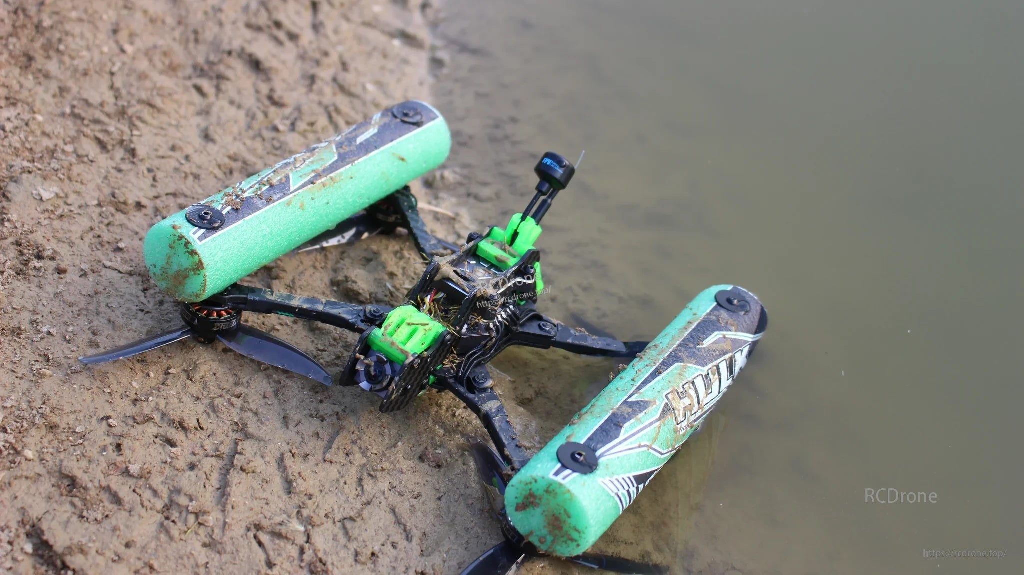 DarwinFPV HULK waterproof FPV drone resting on muddy shoreline beside water, showing rugged outdoor use