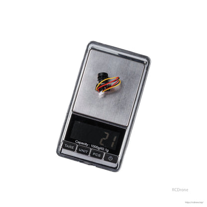 Flywoo 1S Nano Camera, Compact nano FPV camera module with lens and multi-wire connector placed on a digital pocket scale