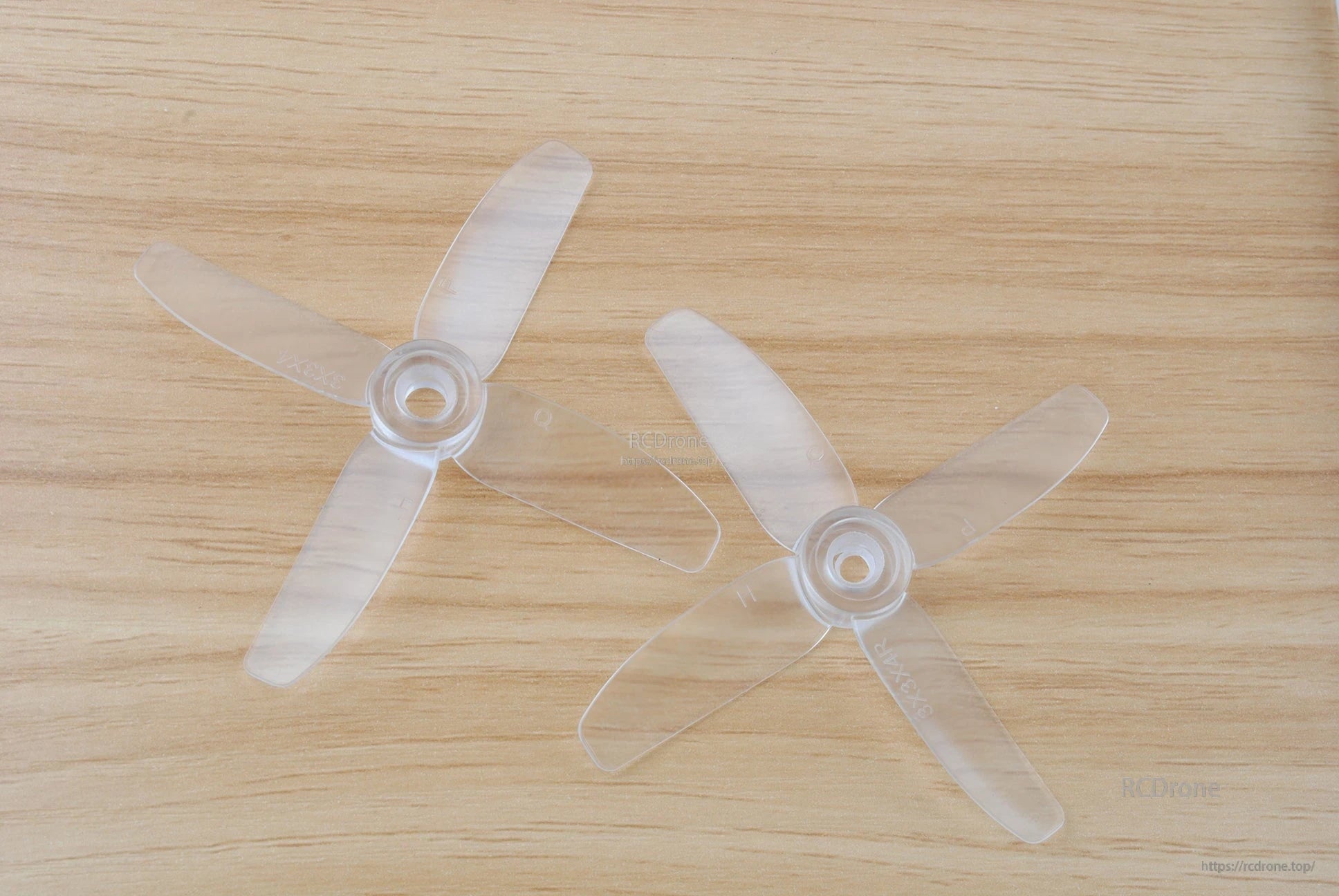 Clear translucent 4-blade propellers pair on wooden surface, top-down view showing hubs and blade shape