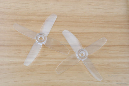 Clear translucent 4-blade propellers pair on wooden surface, top-down view showing hubs and blade shape