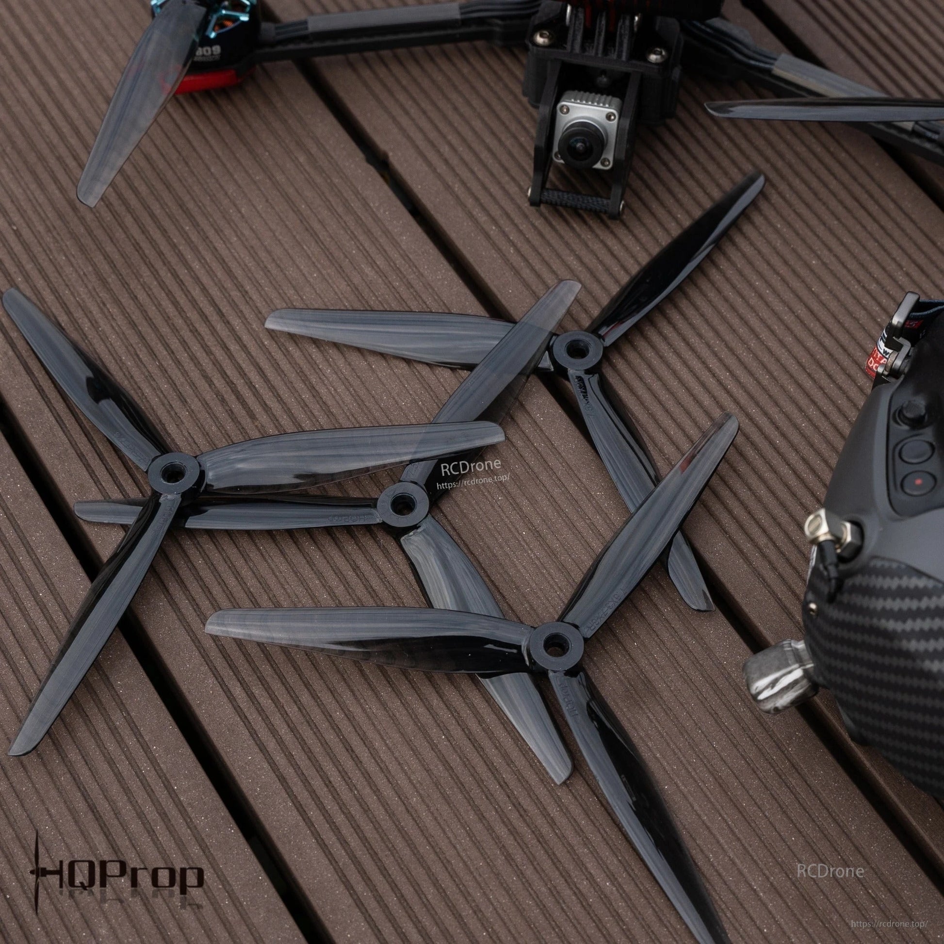 Tri-blade propellers laid on deck next to FPV drone frame and radio transmitter, dark grey finish