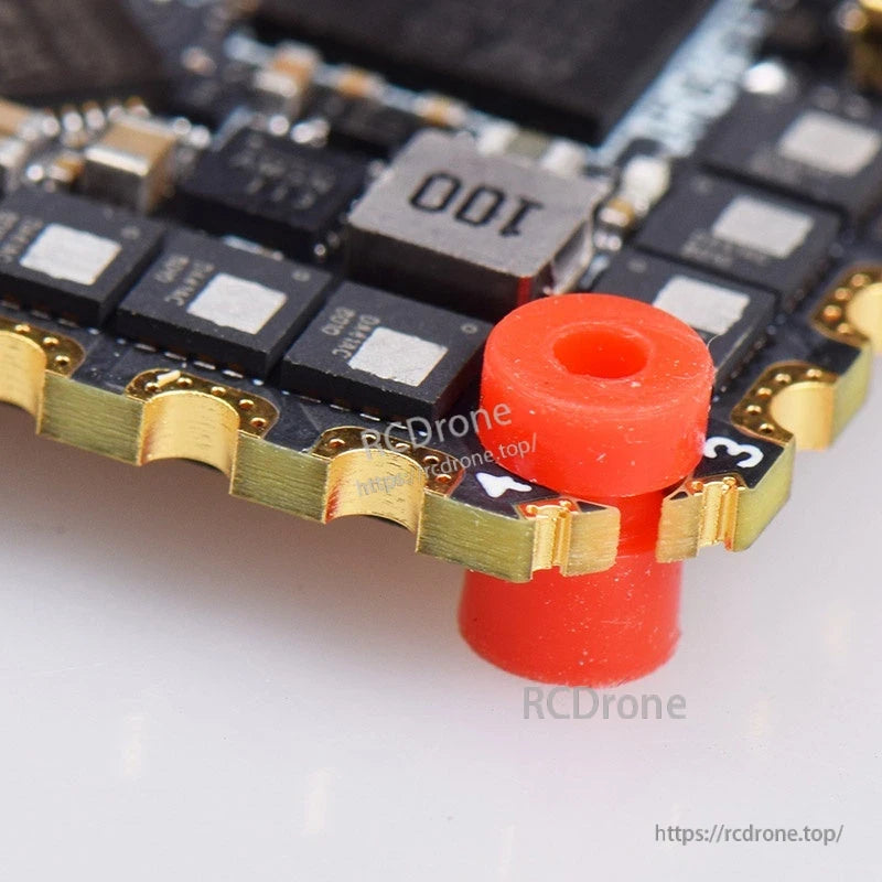 Close-up of PCB corner with red soft mounting grommet, edge solder pads, and 100 inductor on AIO board