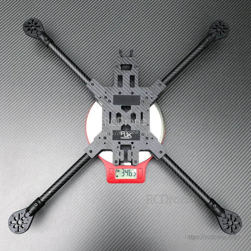 RJX carbon fiber FPV quadcopter frame kit with long arms in X layout, resting on a digital scale