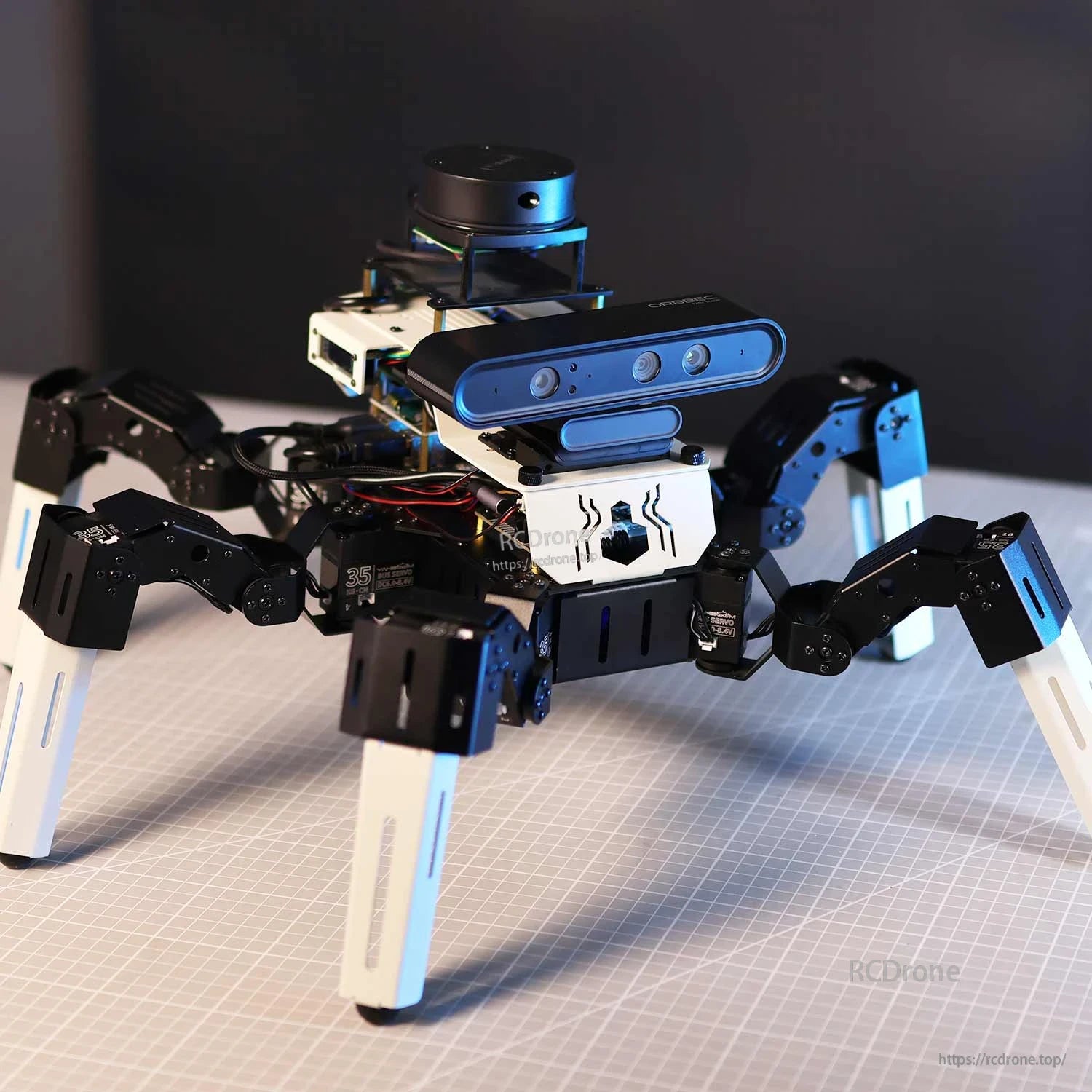 Hexapod robot on cutting mat under studio lighting showing leg joints, wiring, depth camera and top sensor
