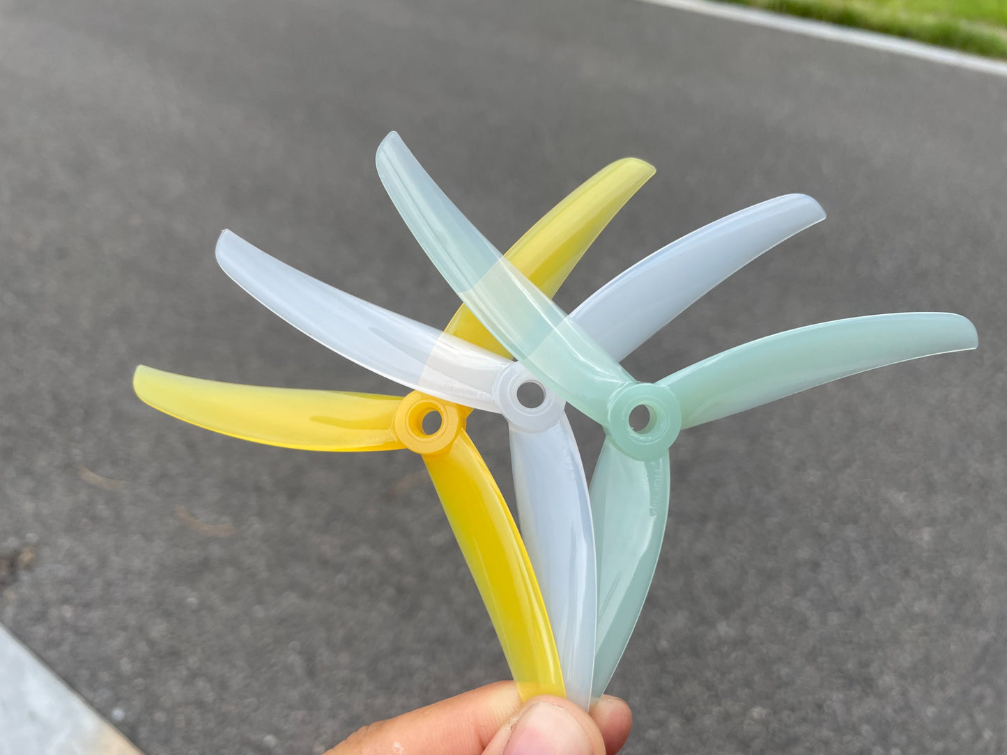 Hand holding three translucent 3-blade FPV drone propellers in yellow, clear, and mint